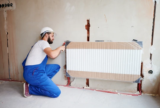 Ouvrier en combinaison bleue et casque blanc installant un radiateur chauffage central dans une pièce en rénovation