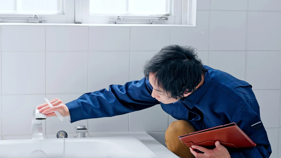 Plombier en combinaison bleue inspecte robinetterie de baignoire blanche avec tablette rouge dans salle de bain carrelée blanc